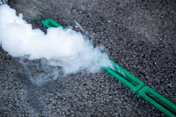 Cloud of fumes disappearing through narrow vents in the road, with green edging.