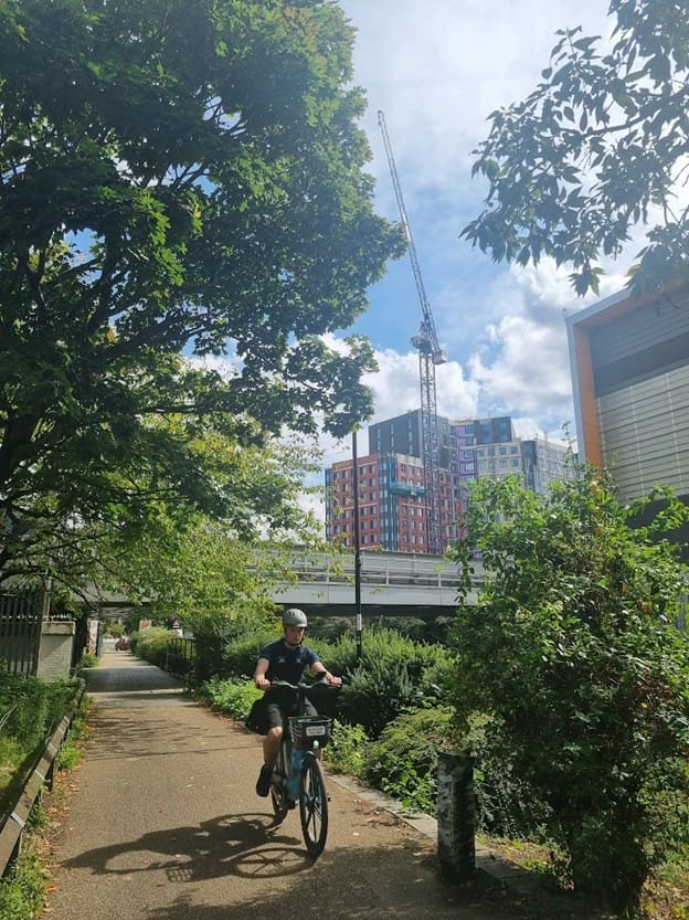 Cyclist heads west on the C10 while new development rises above. In between, the Overground line towards New Cross Gate.