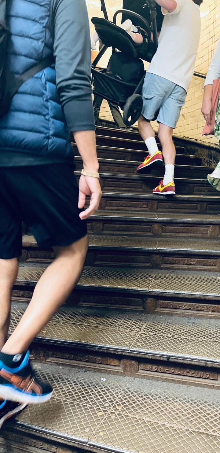 Man carries a buggy containing a small baby up the spiral stairs, other pedestrians seen.