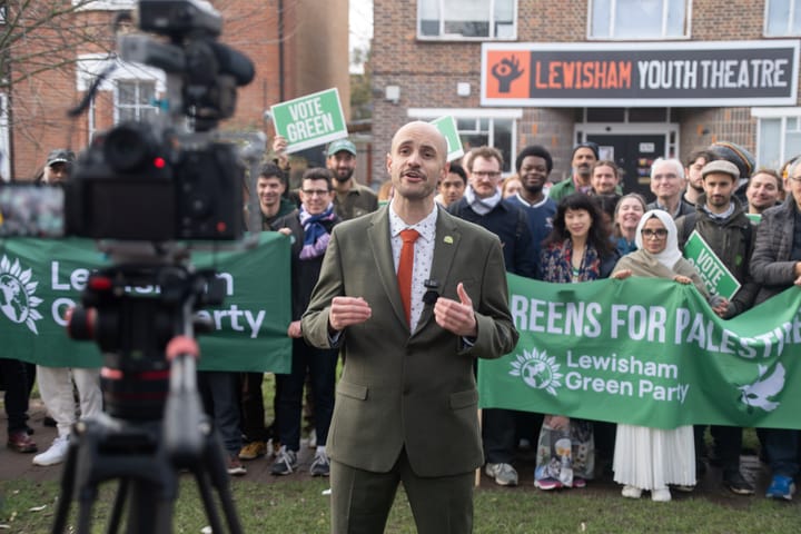 Liam Shrivastava speaks to camera at the launch, supporters behind him with Greens for Palestine banner.